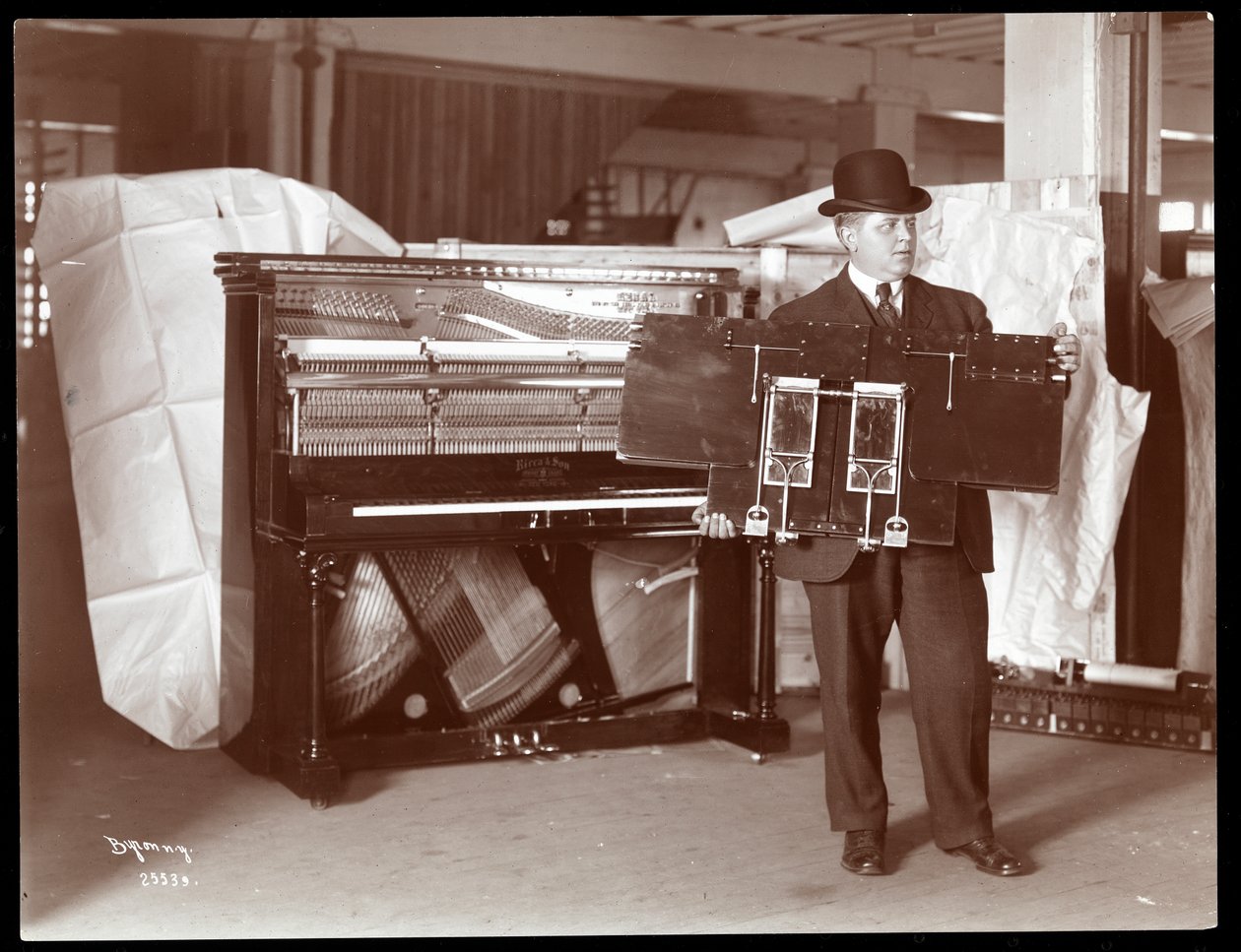 Un piano de Ricca und Son Piano Co. con un hombre adjunto sosteniendo parte de las obras del piano, 1907 de Byron Company
