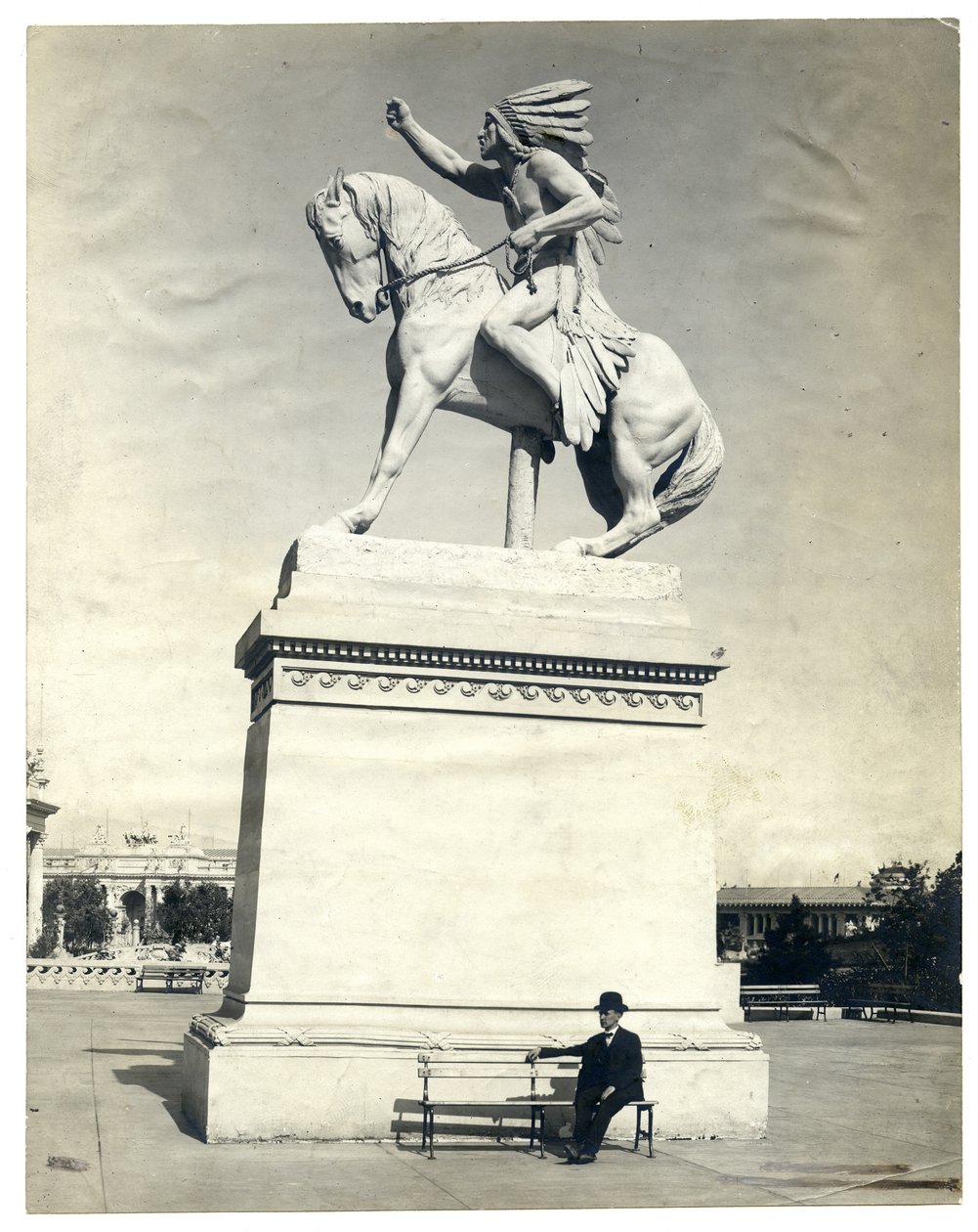 Fotografía vertical en sepia que muestra una estatua de un indio americano sentado en un caballo sobre un gran pedestal. Hay un hombre con traje oscuro y sombrero sentado en un banco en la base del pedestal de Unknown