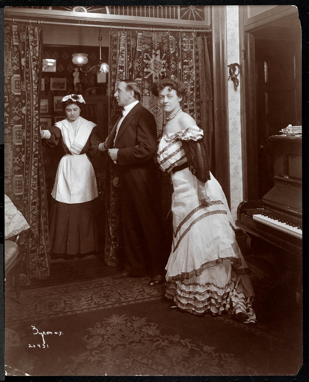 Un hombre y una mujer formalmente vestidos y lo que parece ser un sirviente doméstico en una habitación con un piano, 1905 de Byron Company