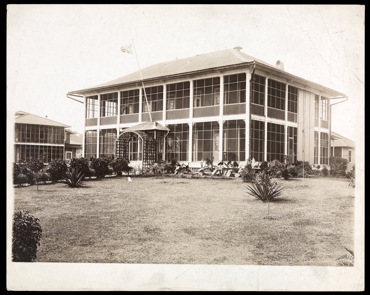Una casa de dos pisos rodeada de porches, con una bandera estadounidense ondeando; en Panamá durante la construcción del canal, 1912 de Byron Company