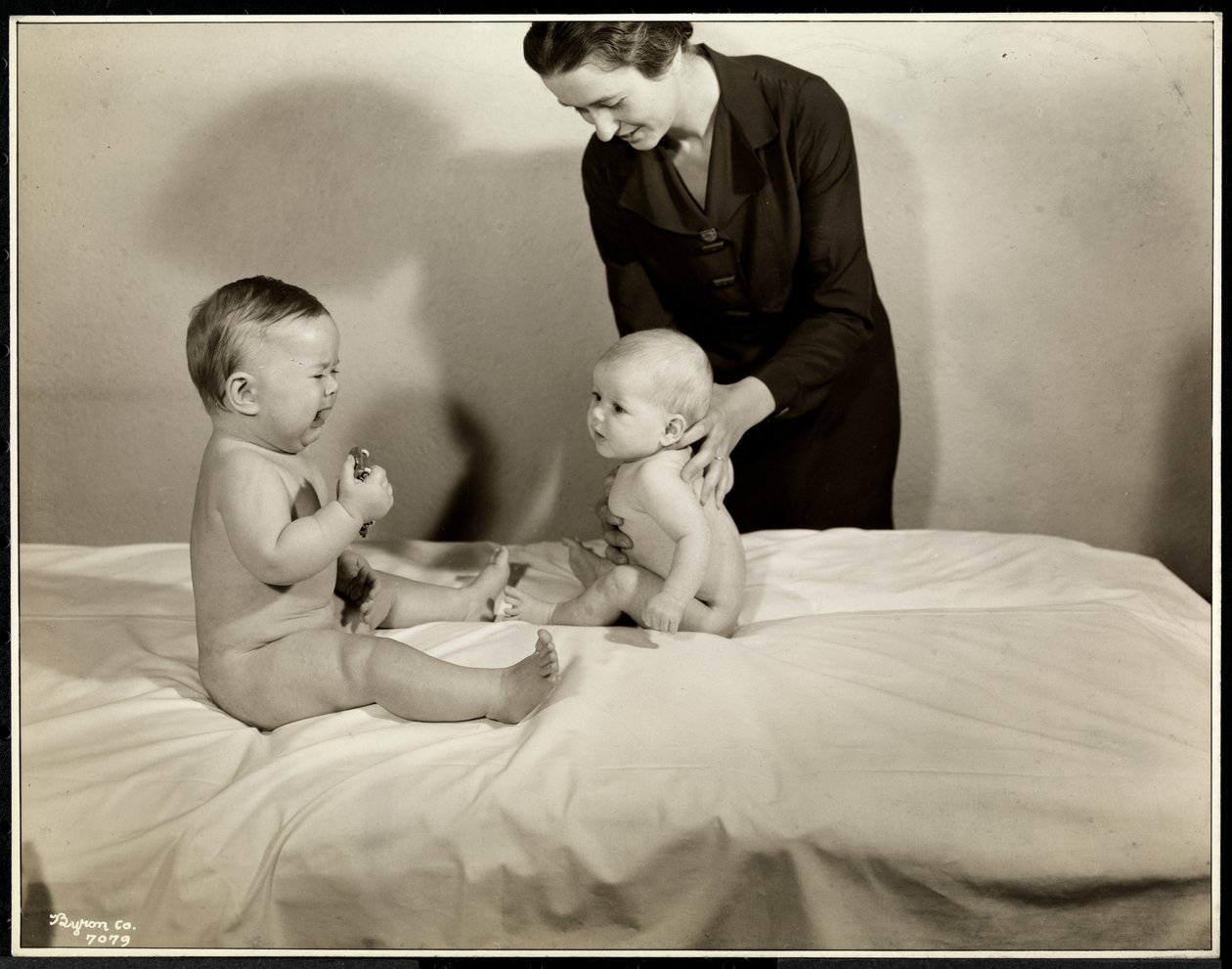 Dos bebés sentados en la cama, uno sostenido por una enfermera, el otro con un coche de juguete y llorando en el Centro de Maternidad en la calle 70 East River, Nueva York, 1935 de Byron Company