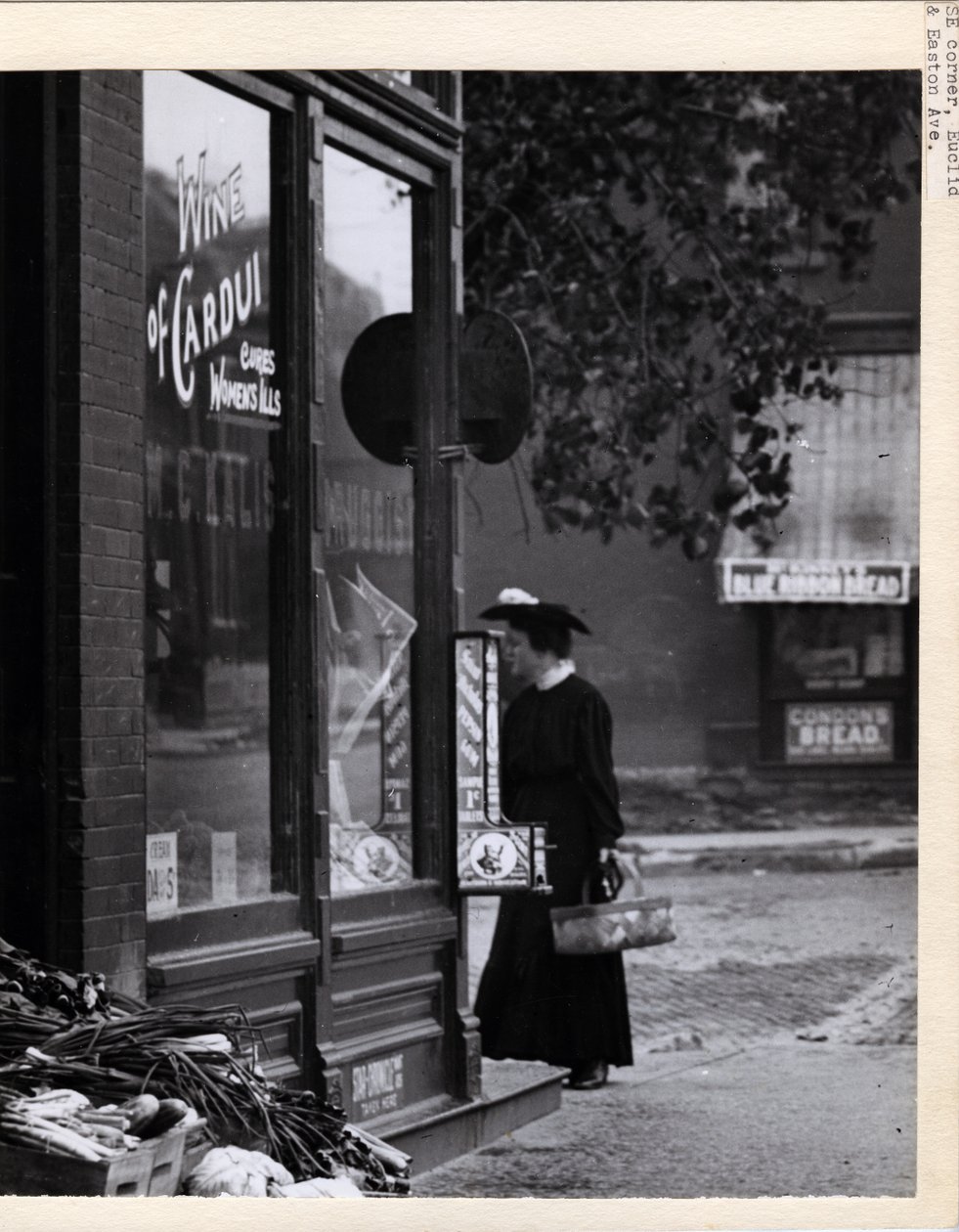 Fotografía vertical en blanco y negro que muestra a una mujer sosteniendo una canasta mientras está de pie frente a la entrada de la farmacia W. C. Kalis en 4878 Easton Avenue de Charles Clement Holt