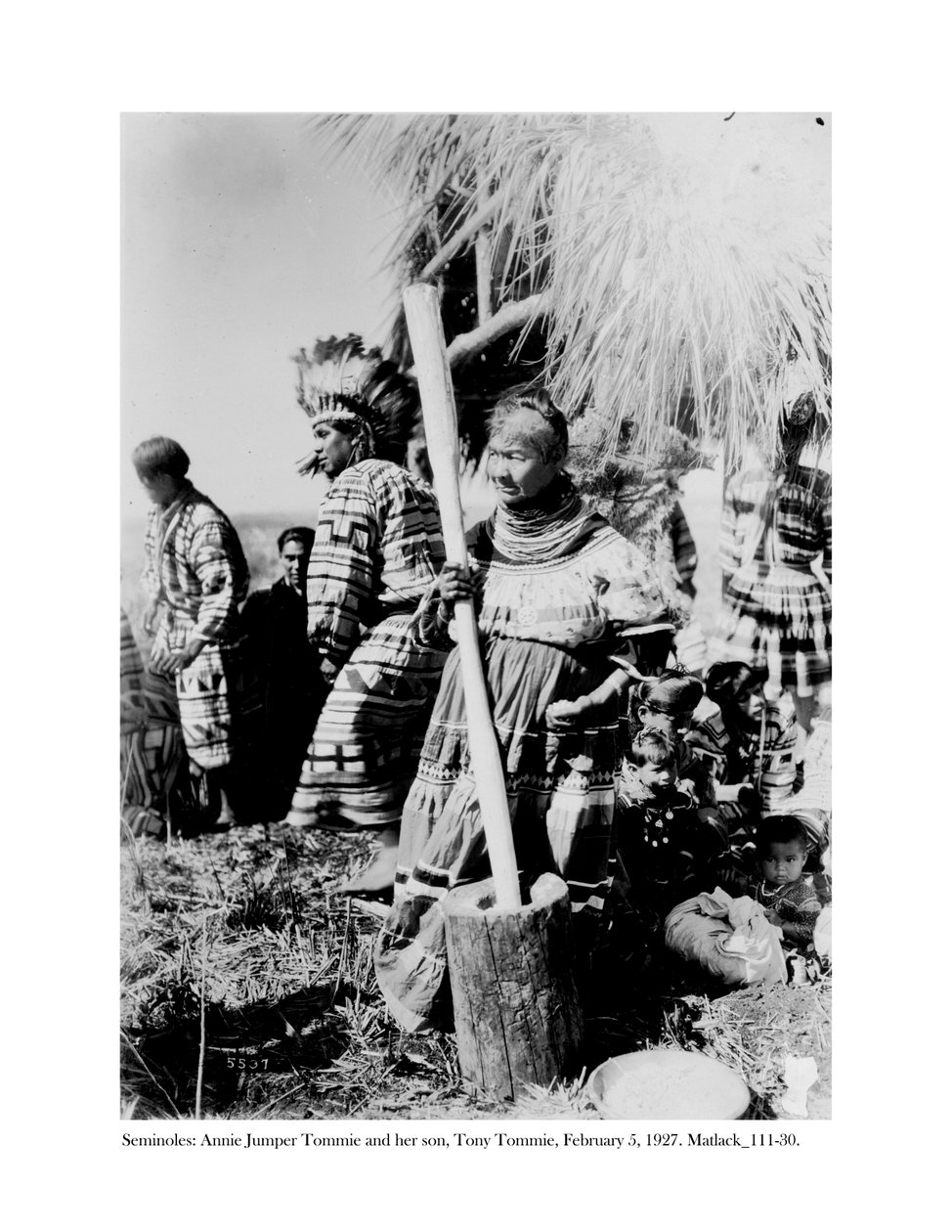 Annie Jumper Tommie usando un mortero y pilón para hacer harina de maíz, y su hijo, Tony Tommie, con un tocado de plumas de águila, 5 de febrero de 1927 de Claude C. Matlack