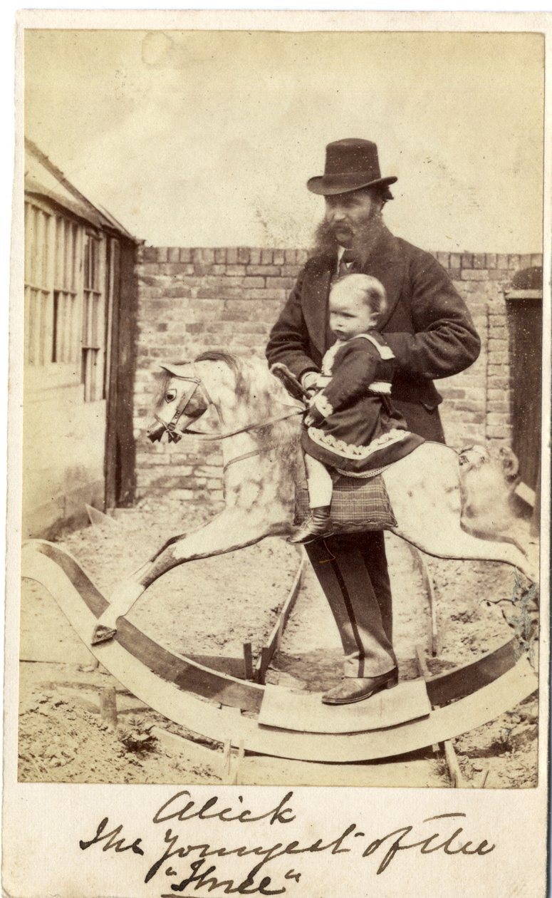Niña en un caballo balancín en el jardín, su padre al lado de English Photographer