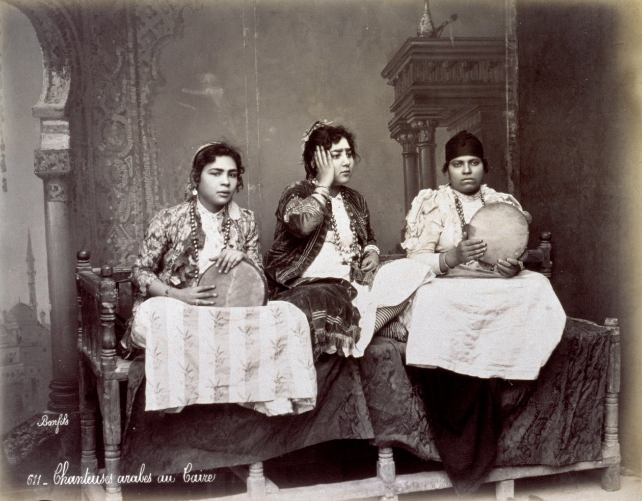 Retrato de tres jóvenes cantantes árabes con vestimenta étnica y collares largos. Están posando en el estudio, sentados. Dos de ellos sostienen una pandereta de Félix Bonfils