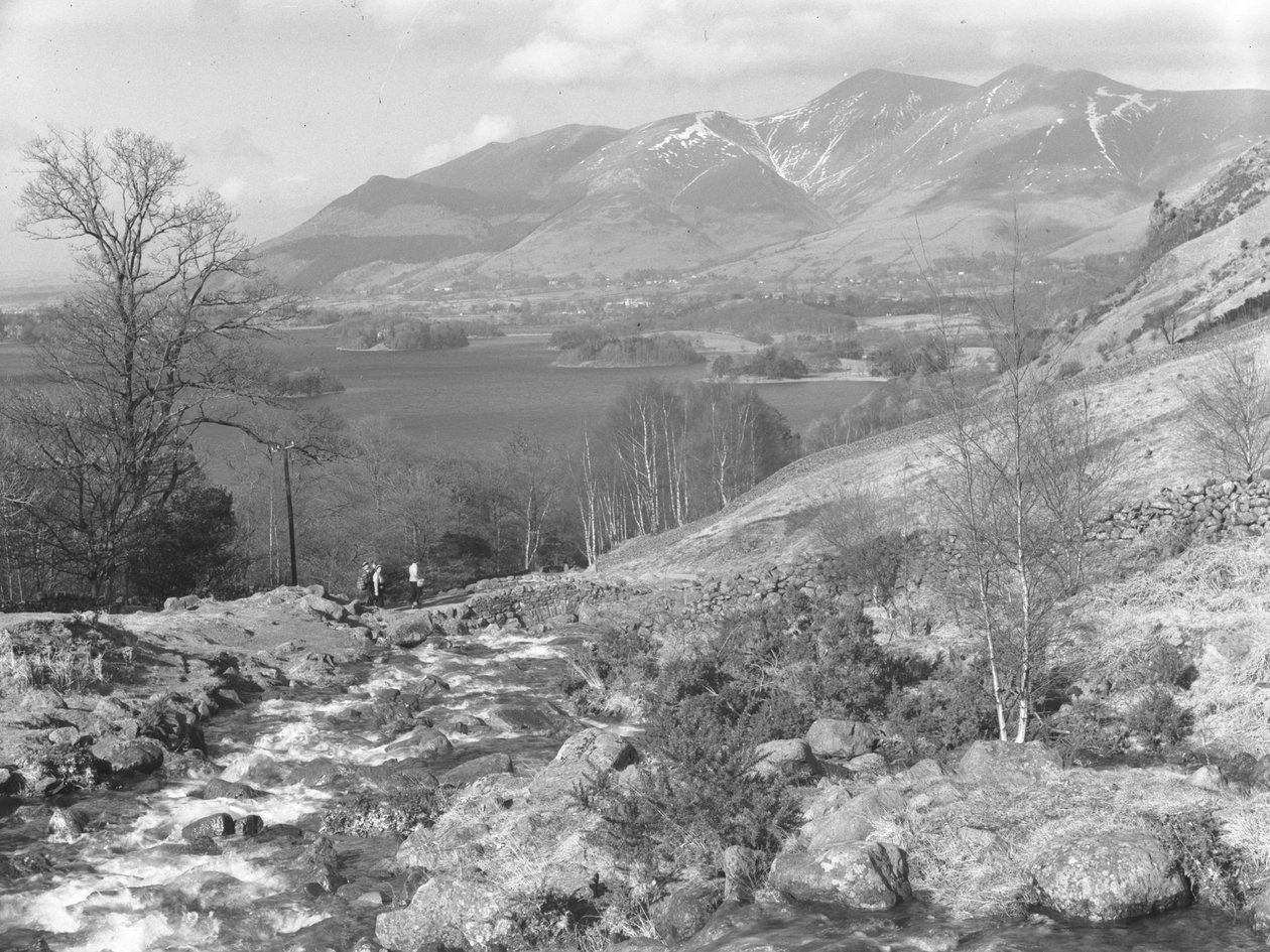 Un grupo de senderistas cruza un puente de piedra seca sobre un arroyo, con las montañas y el lago al fondo. de Joseph Hardman