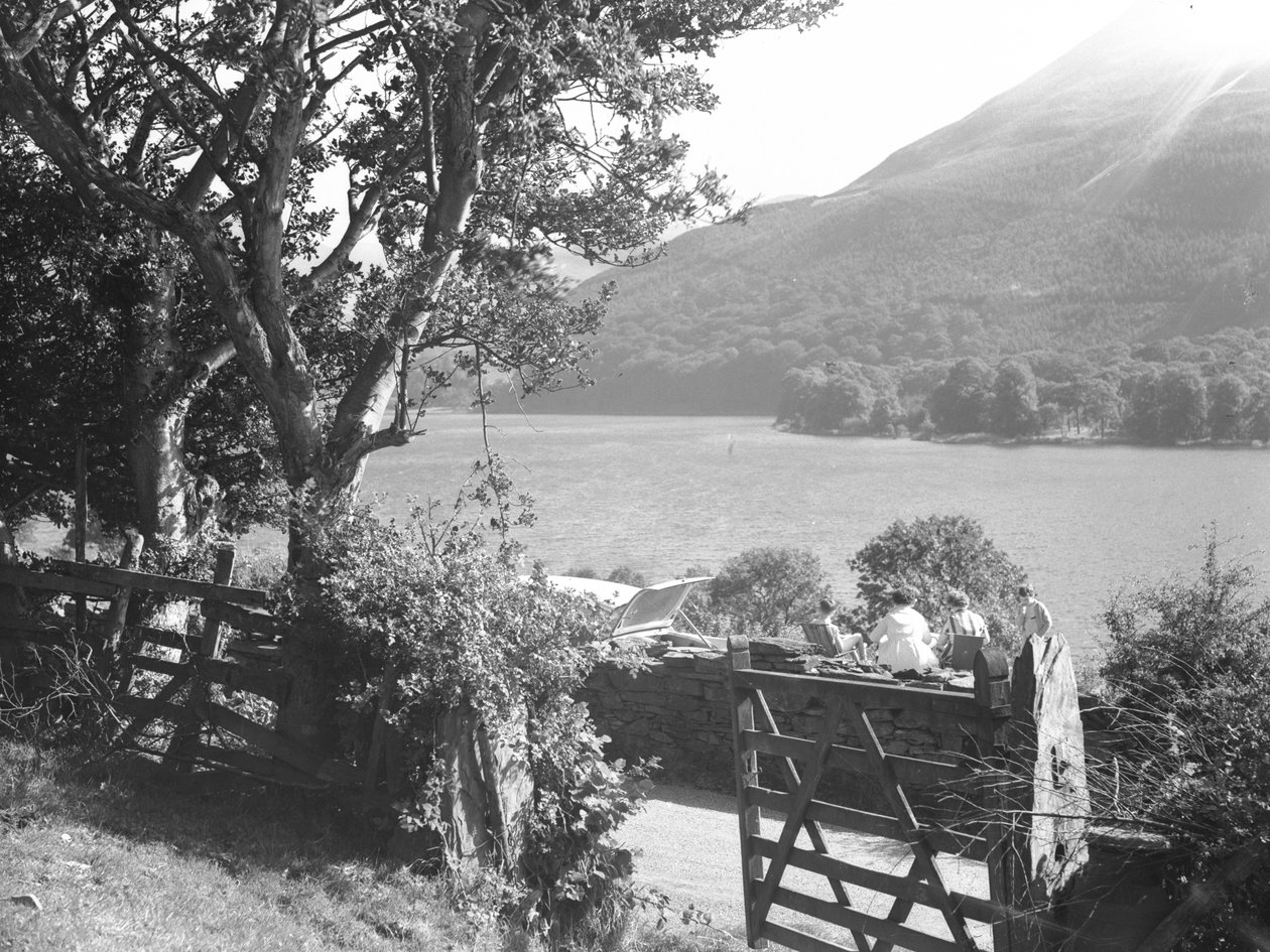 Cuatro personas sentadas en un muro de piedra seca haciendo un picnic con vistas al lago, el coche aparcado con el maletero abierto de Joseph Hardman