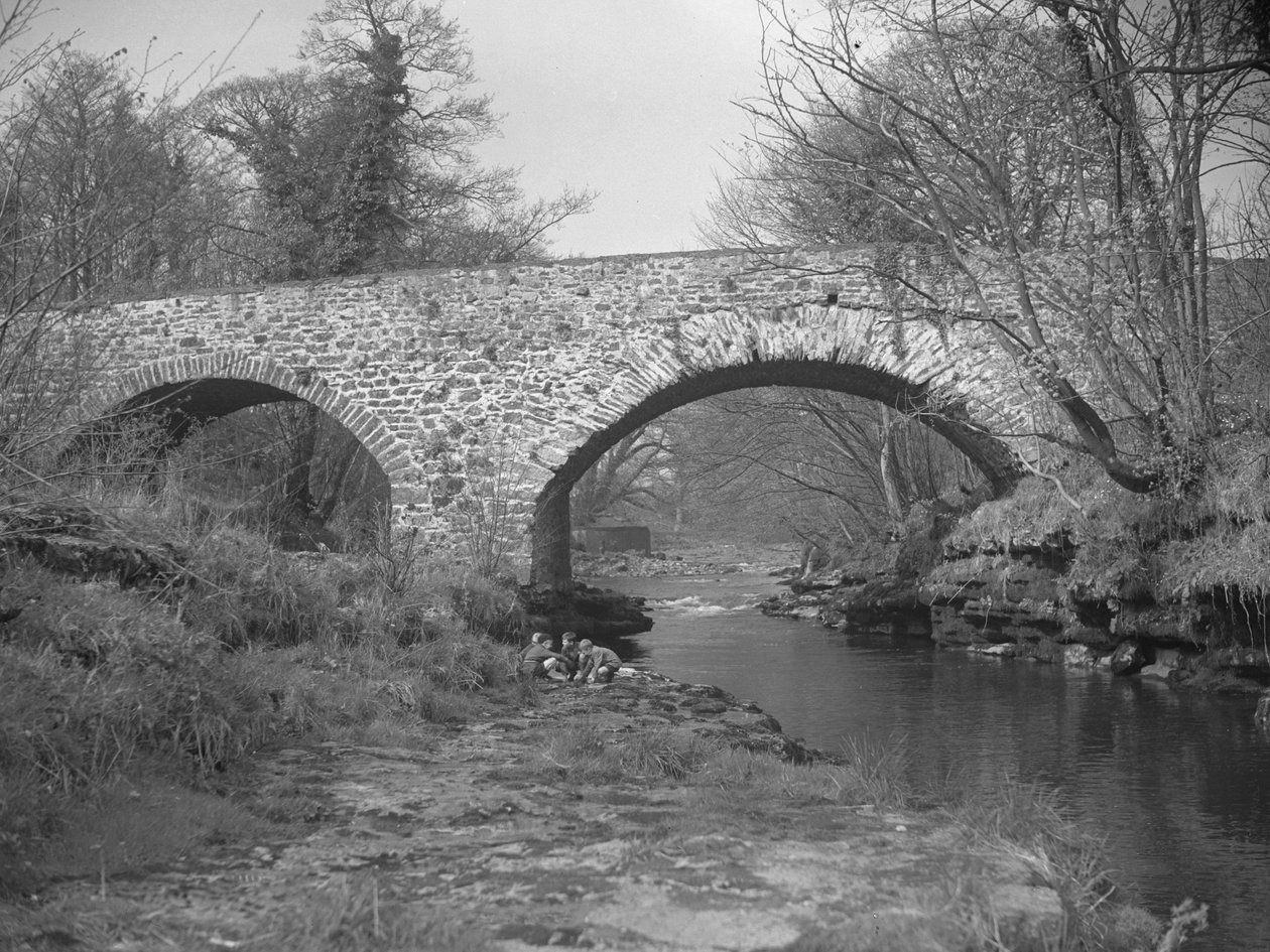 Grupo de chicos agachados en la orilla del río bajo un puente de piedra de Joseph Hardman