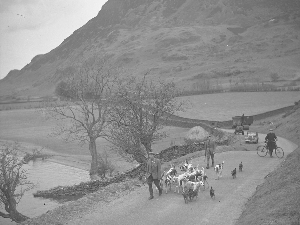 Dos hombres caminando con sabuesos por un carril, detrás hay un hombre con una bicicleta y maquinaria, pajar y lago a la izquierda de la imagen, fells al fondo de Joseph Hardman