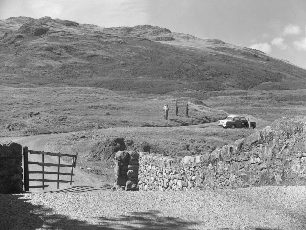 Vista de la carretera que pasa por Hard Knott Pass, dos personas de pie cerca de una señal de tráfico y un coche aparcado en el arcén de hierba. de Joseph Hardman