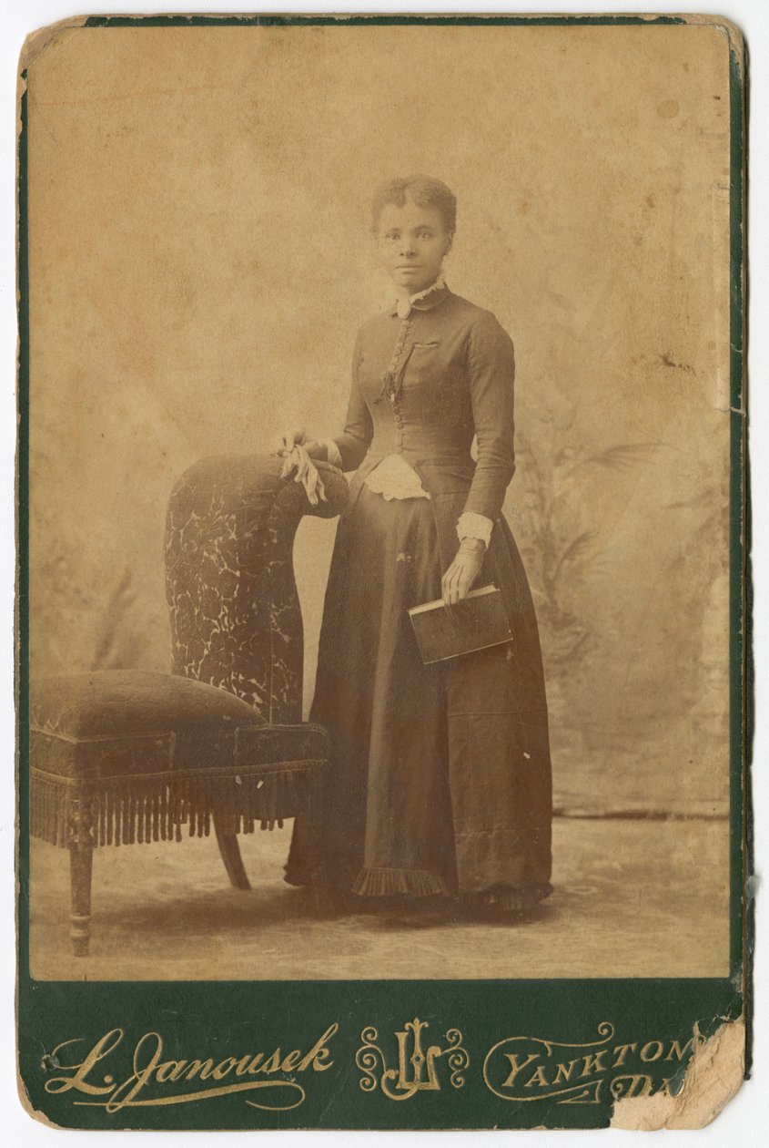 Fotografía de una mujer de pie junto a una silla y sosteniendo un libro, finales del siglo XIX de Louis Janousek