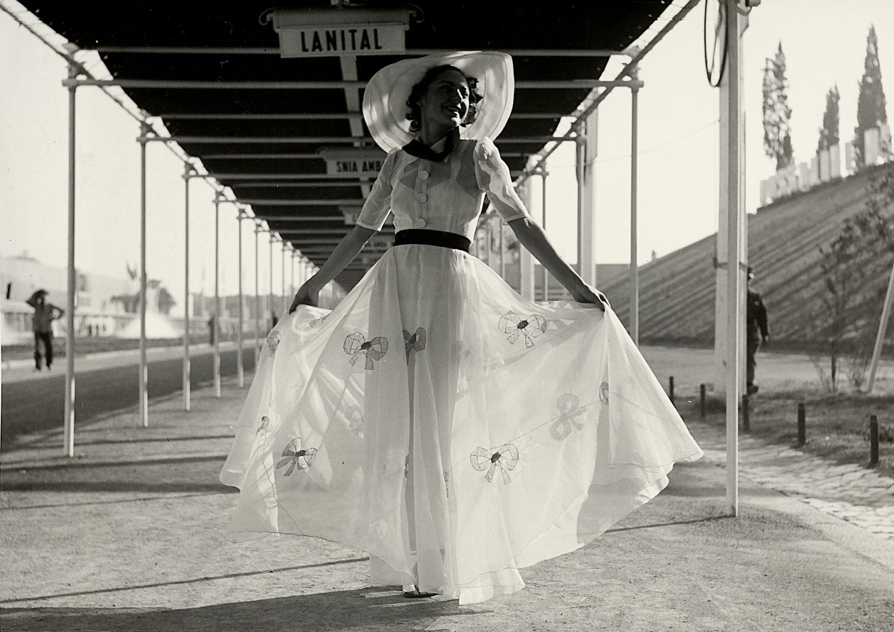 Modelo en un vestido de verano completo creado por la casa de alta costura "Graldi". El vestido es de una tela ligera decorada con bordados; un sombrero de ala ancha completa el conjunto. de Luis Bogino