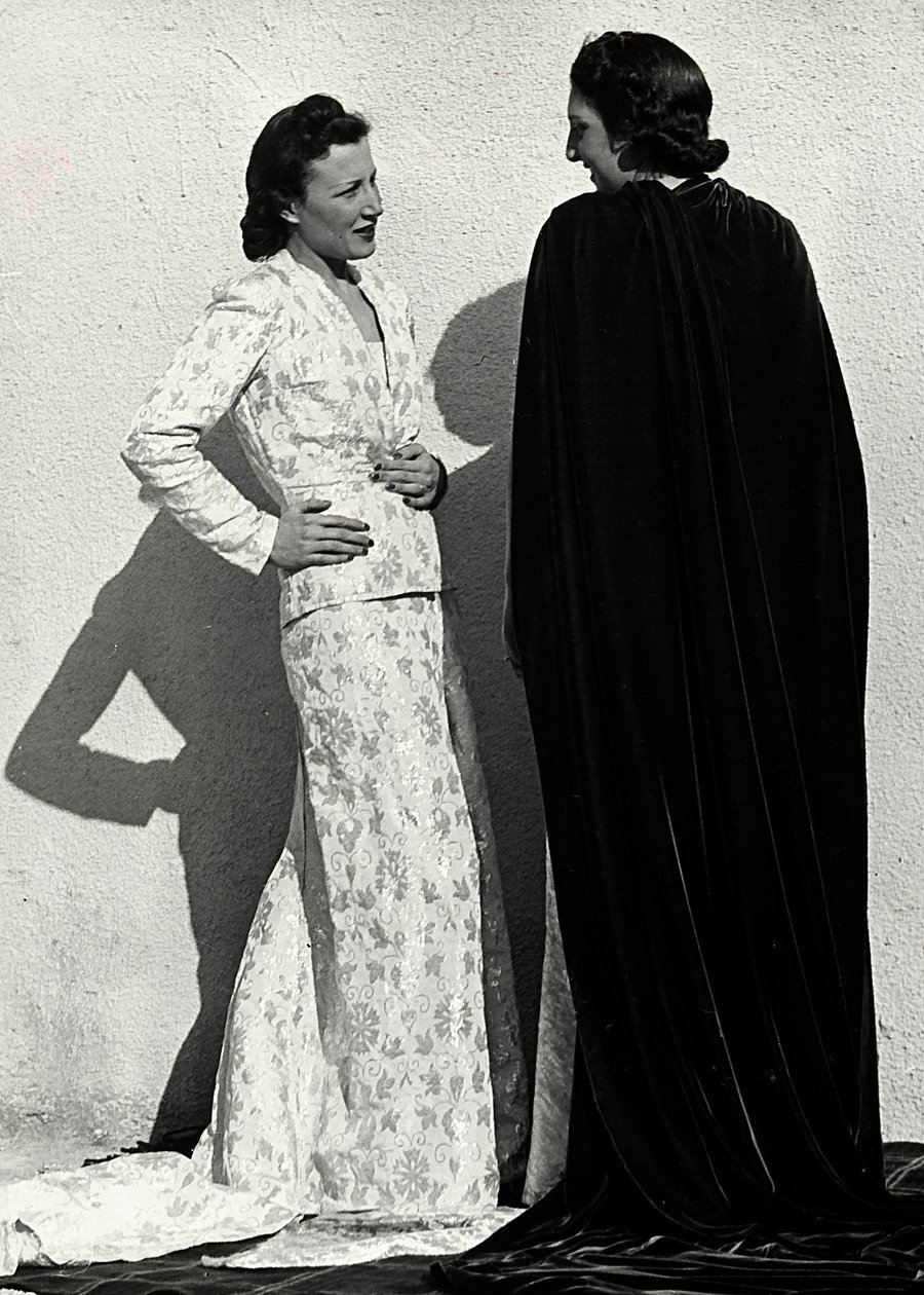 Dos modelos presentan creaciones de la casa de alta costura italiana "Dragoni". Una modelo lleva un conjunto de material estampado compuesto por una chaqueta y una falda larga con cola. La otra modelo, vista desde atrás, lleva un manto largo de terciopelo. de Luis Bogino