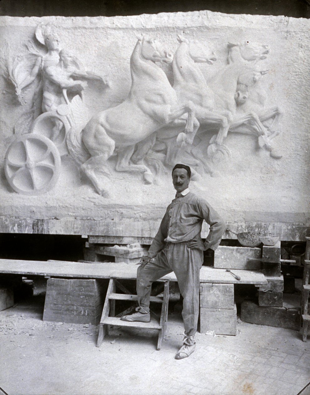 Retrato de cuerpo entero del famoso escultor Pietro Canonica. Está posando en su estudio frente a un alto relieve de mármol en el que está trabajando de Mario Nunes Vais
