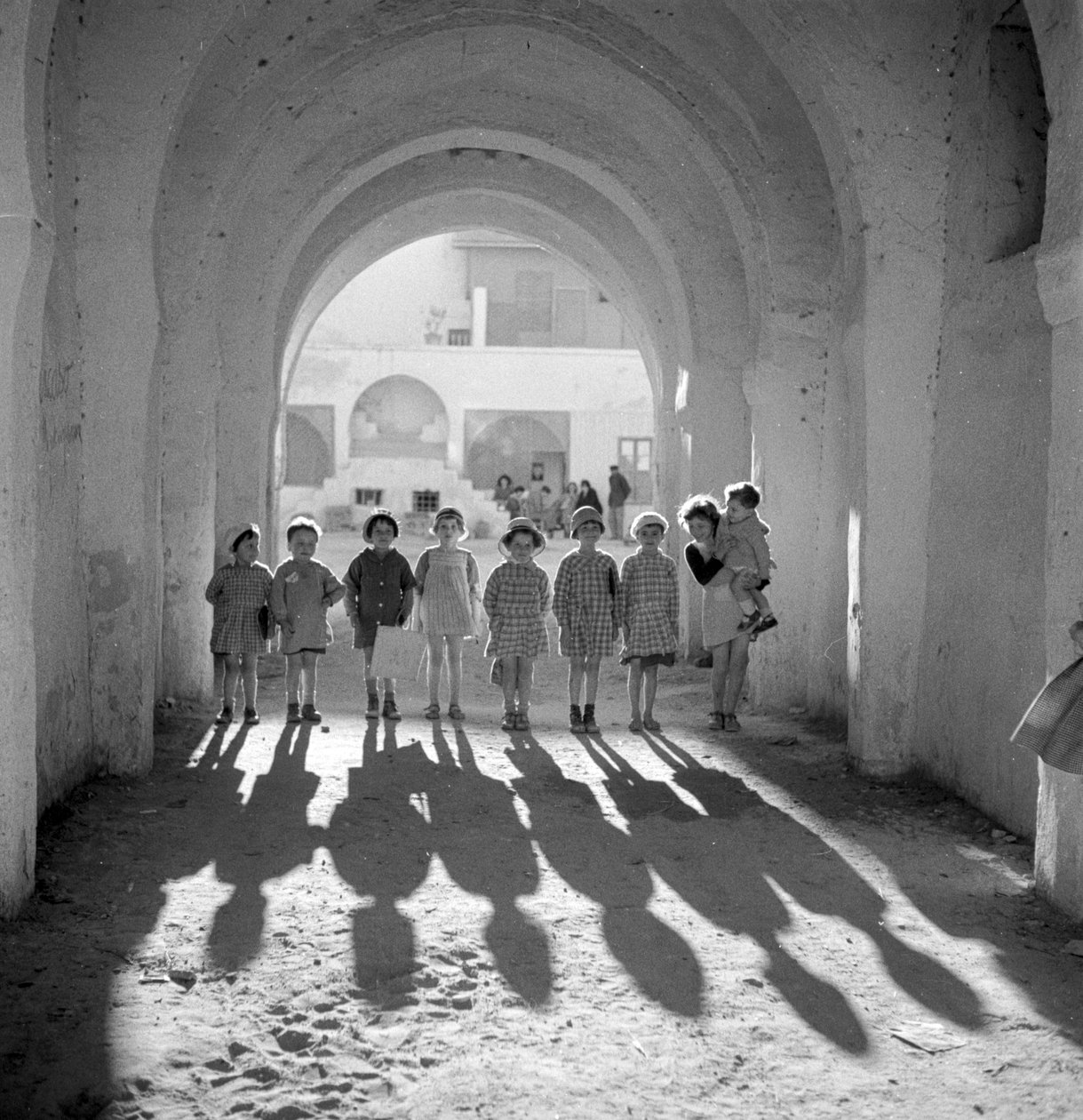 Niños europeos en uniforme escolar bajo arco con sombras. de Maynard Owen Williams