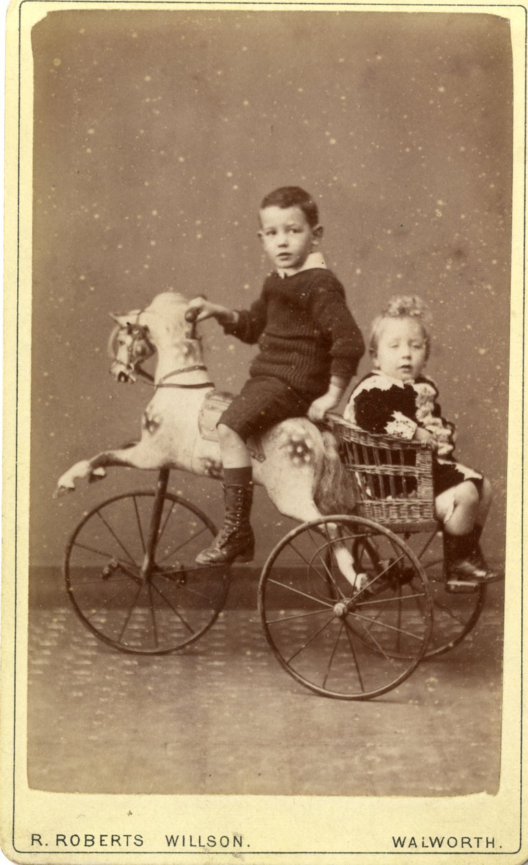 Niño en caballo de juguete con niña en carro de mimbre detrás de R. Roberts Willson