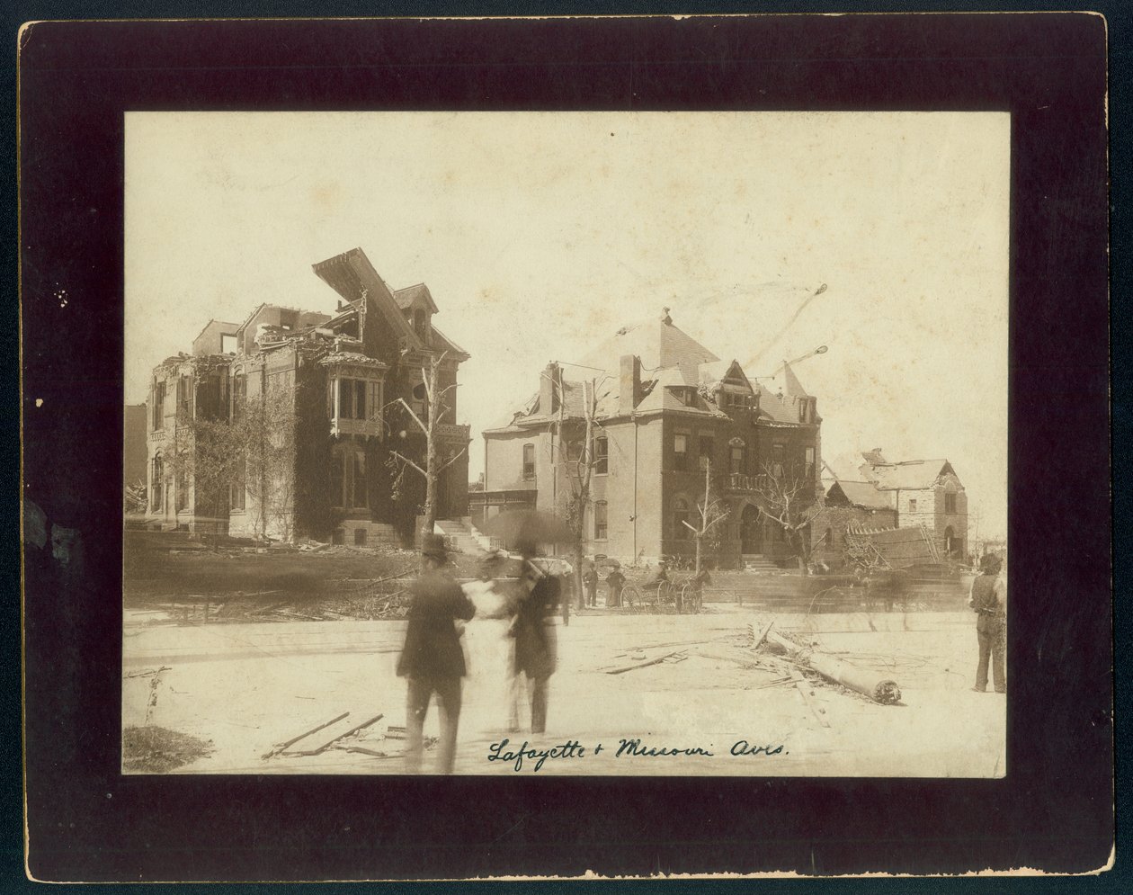 Foto horizontal en sepia mostrando tres casas parcialmente destruidas, árboles despojados y espectadores mirando los daños causados por un tornado de Unbekannt Unbekannt