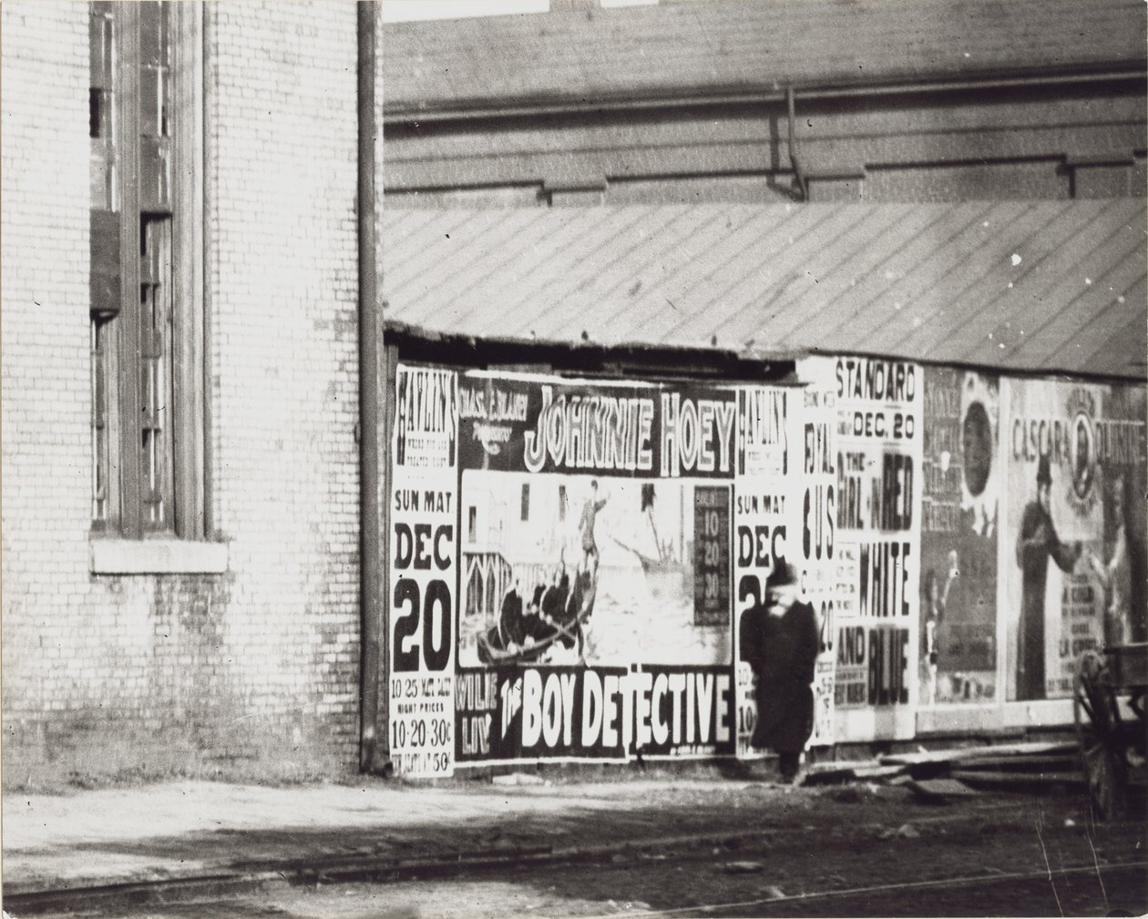Un hombre pasa junto a una pared de anuncios de carteles, incluido uno de Johnnie Hoey, el niño detective, que se presenta en el Teatro Havlin en 600 Walnut Street. de Unbekannt