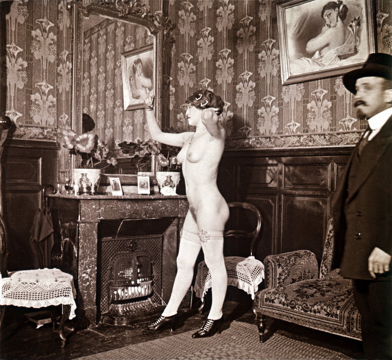 Enigmática fotografía que muestra a una joven completamente desnuda (salvo los zapatos y las medias) peinándose ante un espejo en presencia de un hombre vestido y calentado con un sombrero. Podría pensarse que se trata de una foto tomada en un burdel, pero de Unbekannter Künstler