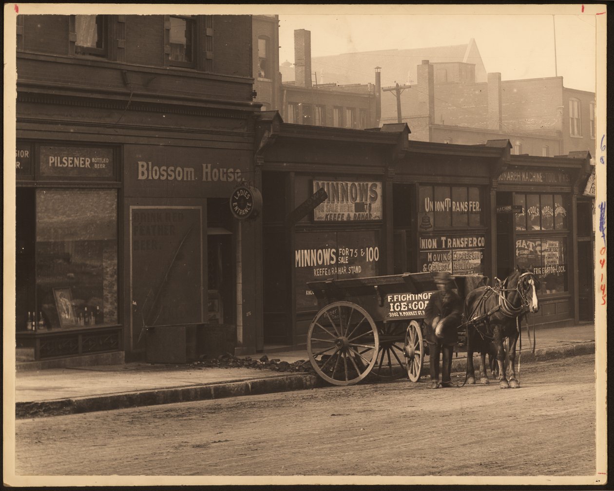 Fotografía horizontal en blanco y negro de una fila de edificios comerciales y un carro tirado por caballos estacionado contra la acera de William G. Swekosky