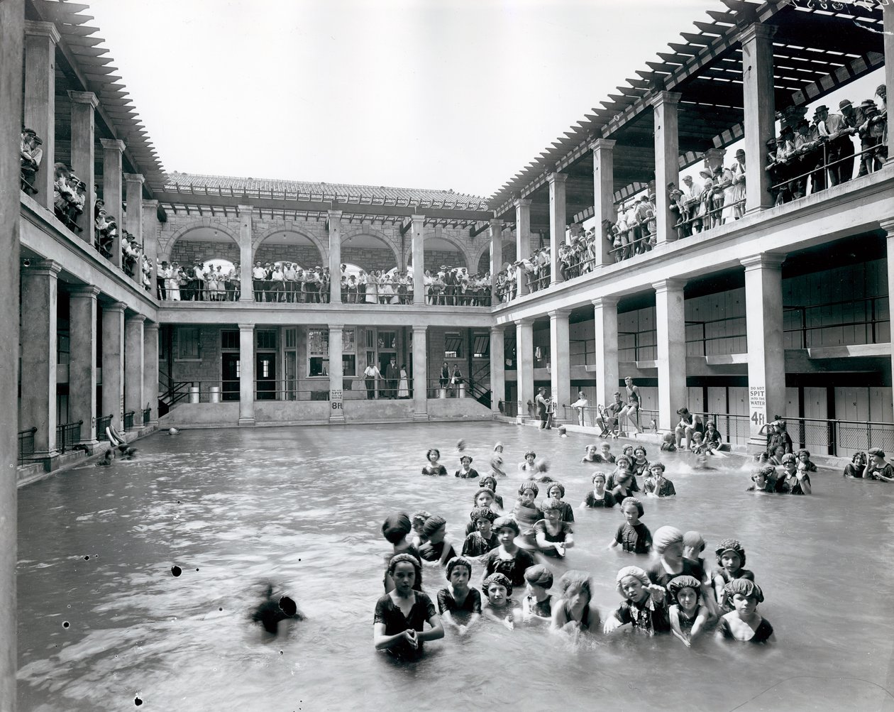 Fotografía horizontal en blanco y negro de una piscina con balcón de dos pisos sostenido por columnas. Espectadores observan a los nadadores desde el balcón del segundo nivel de William G. Swekosky