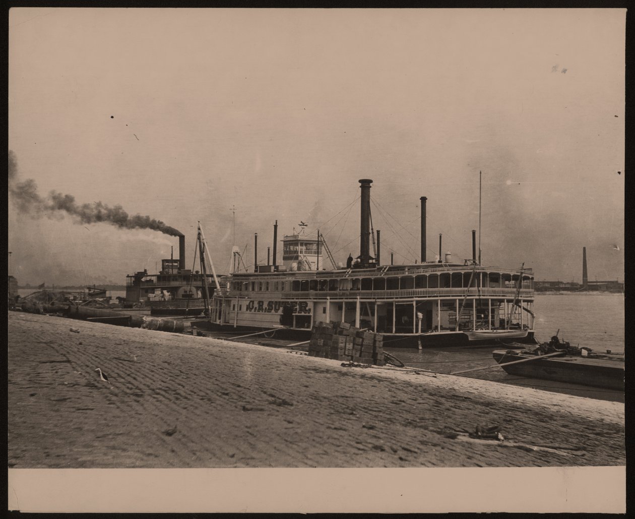 Fotografía horizontal en blanco y negro del barco de vapor C.R. Suter. Otro barco de vapor está estacionado detrás y hay cajas apiladas en la orilla de William G. Swekosky