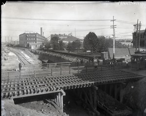 Fotografía horizontal en blanco y negro que muestra un puente de madera y vías de tranvía sobre vías de ferrocarril en construcción. Un tranvía marcado Chouteau 2137 está comenzando a cruzar el puente a la derecha. En la distancia, edificios de fábricas co de Charles Clement Holt