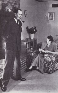 Esposa lee, mientras el esposo calienta su espalda contra el fuego, c.1950