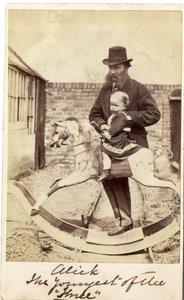 Niña en un caballo balancín en el jardín, su padre al lado de English Photographer