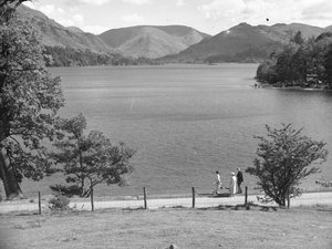 Un hombre, una mujer y posiblemente su hija pasean por la orilla de un lago, con unas colinas al fondo.