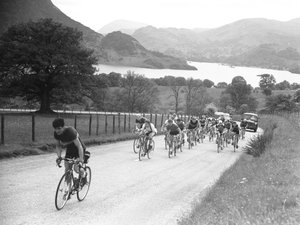 Grupo de ciclistas subiendo por una carretera seguidos de coches, lago y colinas al fondo.