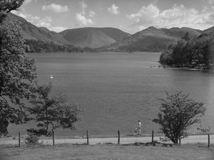 En primer plano, una señora mira hacia un lago; a la derecha, hay coches aparcados y gente jugando en el agua; al fondo, unas colinas.
