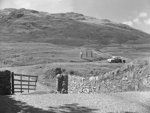 Vista de la carretera que pasa por Hard Knott Pass, dos personas de pie cerca de una señal de tráfico y un coche aparcado en el arcén de hierba. de Joseph Hardman