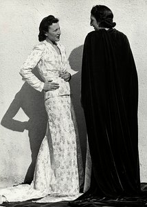 Dos modelos presentan creaciones de la casa de alta costura italiana "Dragoni". Una modelo lleva un conjunto de material estampado compuesto por una chaqueta y una falda larga con cola. La otra modelo, vista desde atrás, lleva un manto largo de terciopelo. de Luis Bogino