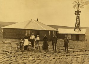 Familia de colonos, fuera de su casa de tepes con un molino de viento en el tejado del edificio contiguo en el condado de Custer, Nebraska, hacia 1885 (fotografía en blanco y negro). de Solomon D. Butcher