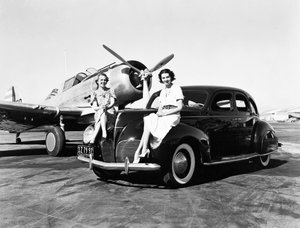 California: 1939 Dos mujeres sentadas en los guardabarros de un Lincoln Zephyr de 1939 en un aeropuerto con un avión de un solo motor estacionado detrás de ellas (foto en b/n)