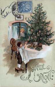 Tarjeta de felicitación navideña: un niño frente a un árbol de Navidad posa sobre una mesa, observado por un ángel en la ventana - Postal fechada en 1905