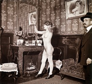 Enigmática fotografía que muestra a una joven completamente desnuda (salvo los zapatos y las medias) peinándose ante un espejo en presencia de un hombre vestido y calentado con un sombrero. Podría pensarse que se trata de una foto tomada en un burdel, pero de Unbekannter Künstler
