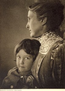 Retrato de medio cuerpo de un niño tiernamente abrazado por una anciana. La mujer posa de perfil, sosteniendo el pecho del pequeño. Tiene una mirada vivaz y pícara, que dirige hacia la cámara.