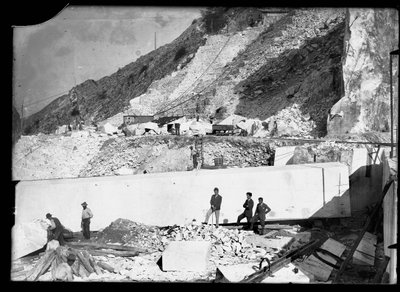 Tres hombres observan el trabajo de varias excavadoras en los Alpes Apuanos. Detrás de ellos hay un enorme bloque de mármol ya cuadrado y listo para ser transportado al valle (foto en b/n) de A. Corsini