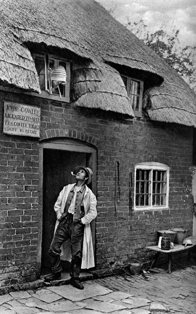 Un hombre fumando una pipa fuera de una tienda, Worcestershire, c1922 de A. W. Cutler