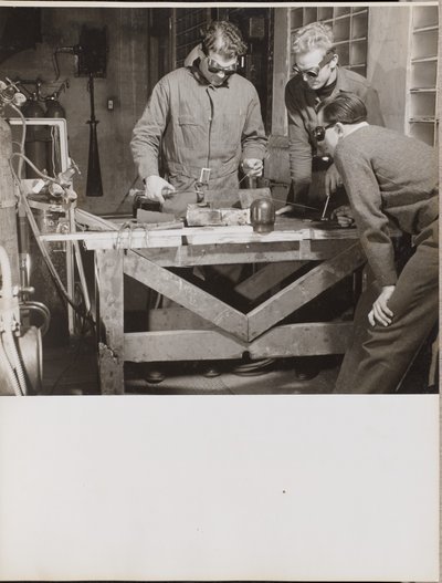 [Tres hombres con gafas sueldan una pieza de metal sobre una mesa] de Arnold Eagle