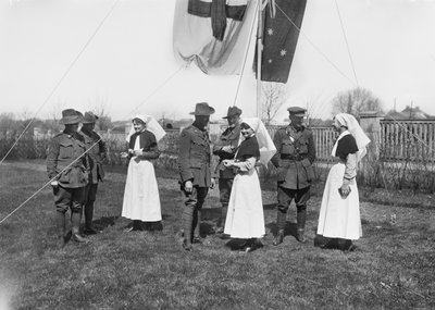 Un grupo de oficiales, enfermeras y hombres no identificados de la 3ª Estación de Despeje de Bajas Australiana reunidos bajo un asta de bandera ondeando la bandera australiana (Australian Blue Ensign) y la bandera de la Cruz Roja de Australian Photographer