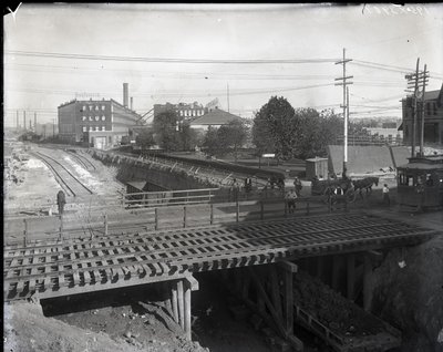 Fotografía horizontal en blanco y negro que muestra un puente de madera y vías de tranvía sobre vías de ferrocarril en construcción. Un tranvía marcado Chouteau 2137 está comenzando a cruzar el puente a la derecha. En la distancia, edificios de fábricas co de Charles Clement Holt