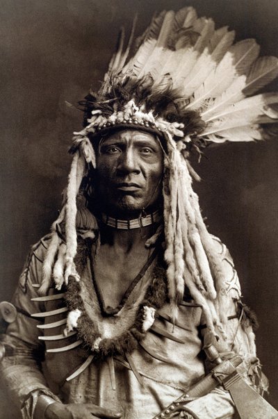 Cola de Comadreja (Apohsuyis). Retrato de jefe indio de la tribu piegan que lleva el famoso tocado de plumas de águila y de comadreja, una túnica de ante, dos collares, uno de hueso y otro de garras de oso pardo, y una pipa tomahawk. de Edward Sheriff Curtis