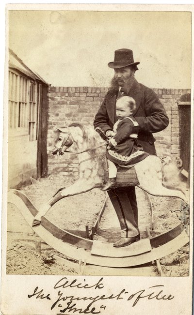 Niña en un caballo balancín en el jardín, su padre al lado de English Photographer