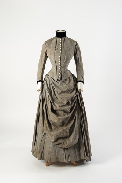 Vestido de algodón gris con delantal drapeado al frente y adornos de terciopelo negro en el cuello y los puños, finales de 1880 de English School