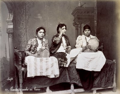 Retrato de tres jóvenes cantantes árabes con vestimenta étnica y collares largos. Están posando en el estudio, sentados. Dos de ellos sostienen una pandereta de Félix Bonfils