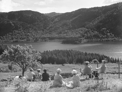 Grupo de personas sentadas en la hierba o en tumbonas mirando una vista de Thirlmere de Joseph Hardman