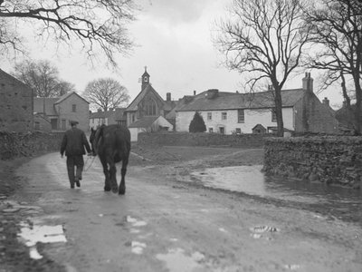 Hombre conduciendo caballo por la carretera lejos de la cámara hacia los edificios del pueblo y la iglesia; arquitectura; religiosa; eclesiástica; de Joseph Hardman
